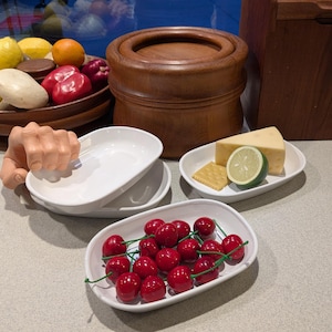 May include: A wooden bowl filled with lemons, oranges, mushrooms, and red peppers. White oval serving dishes hold cherries, cheese, and a lime. A wooden container and a hand-shaped holder for the dishes are also present.