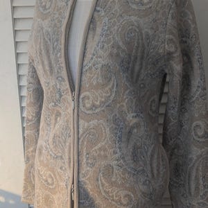 May include: A beige zip-up jacket with a paisley pattern in shades of beige and blue. The jacket has a high collar and a full-length zipper. The jacket is displayed on a mannequin. The jacket is made of a soft, textured material.