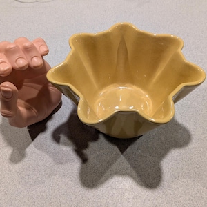 May include: A yellow ceramic bowl with a scalloped edge sits next to a pink hand sculpture. The bowl has a glossy finish and a star-like shape. The hand sculpture is in a fist shape.