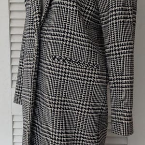 Puede incluir: Un abrigo largo con estampado de pata de gallo en blanco y negro. El abrigo presenta un cuello con muescas, dos bolsillos delanteros y mangas largas. El abrigo se exhibe en un maniqu&iacute; sobre un fondo blanco.