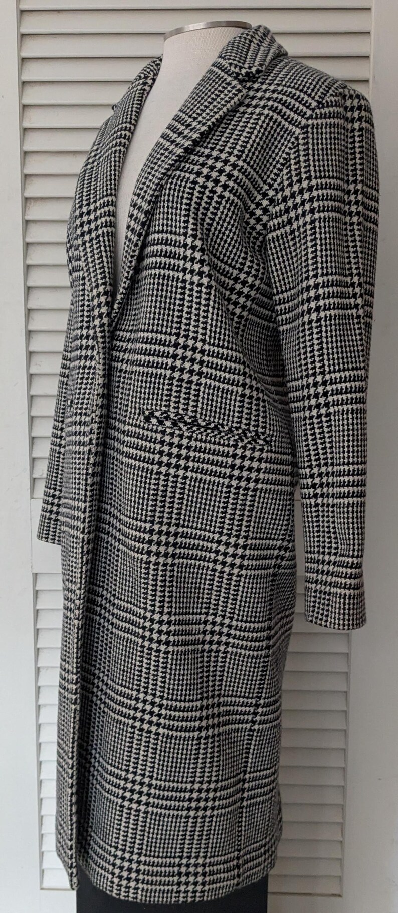 Puede incluir: Un abrigo largo con estampado de pata de gallo en blanco y negro. El abrigo presenta un cuello con muescas, dos bolsillos delanteros y mangas largas. El abrigo se exhibe en un maniqu&iacute; sobre un fondo blanco.