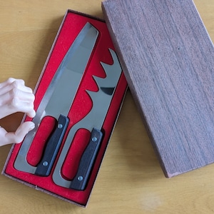 May include: Two silver kitchen knives with black handles are displayed in a red velvet box. The box is open and the knives are visible.