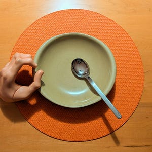 May include: A light beige ceramic bowl with a smooth interior surface and a silver-toned metal spoon resting inside. A pale, realistic-looking plastic hand model is positioned on the left side of the bowl, forming a circular gesture with its fingers.