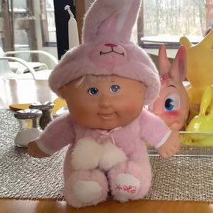 May include: A pink plush Cabbage Patch Kid doll wearing a pink bunny costume with white fur trim. The doll has blonde hair and blue eyes. The doll is sitting on a woven surface.