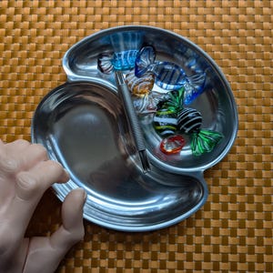 May include: A silver-colored, two-sectioned dish with a decorative, swirled shape. The dish contains several colorful, glass-blown candies in various colors, including blue, green, and orange. A silver utensil rests in the dish. The dish sits on a woven, brown surface.