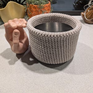 May include: A beige macrame basket with a cylindrical shape. The basket has a thick, textured weave and a dark interior. A ceramic hand sculpture is next to the basket.