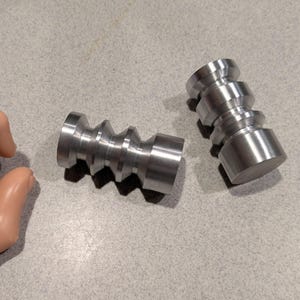 May include: Two cylindrical metal objects with a series of ridges and flat ends. The objects are silver in color and appear to be machined from solid metal. They are lying on a light gray surface.