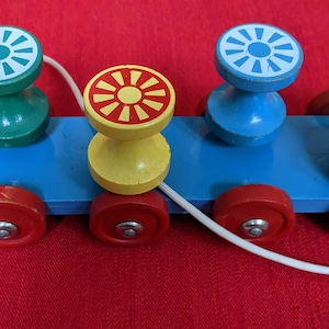 May include: A wooden toy train with four colorful cars. Each car has a different colored top with a sun design. The train has red wheels and a blue base. A white string is attached to the front of the train.