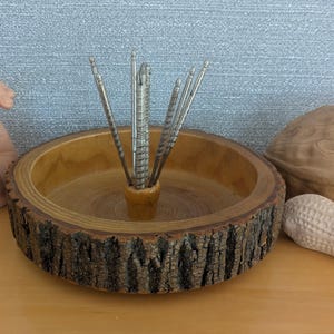 May include: A wooden nut bowl with a bark-like edge, holding silver nutcrackers. A walnut and a peanut are next to the bowl. The bowl has a natural wood grain finish, and the nutcrackers are arranged in the center.