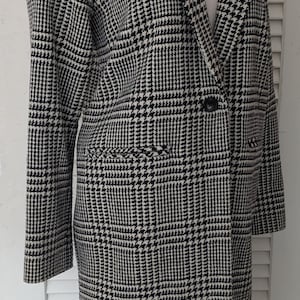 May include: A black and white houndstooth pattern coat with a notched collar and two front pockets. The coat is buttoned and has long sleeves. The coat is displayed on a mannequin.