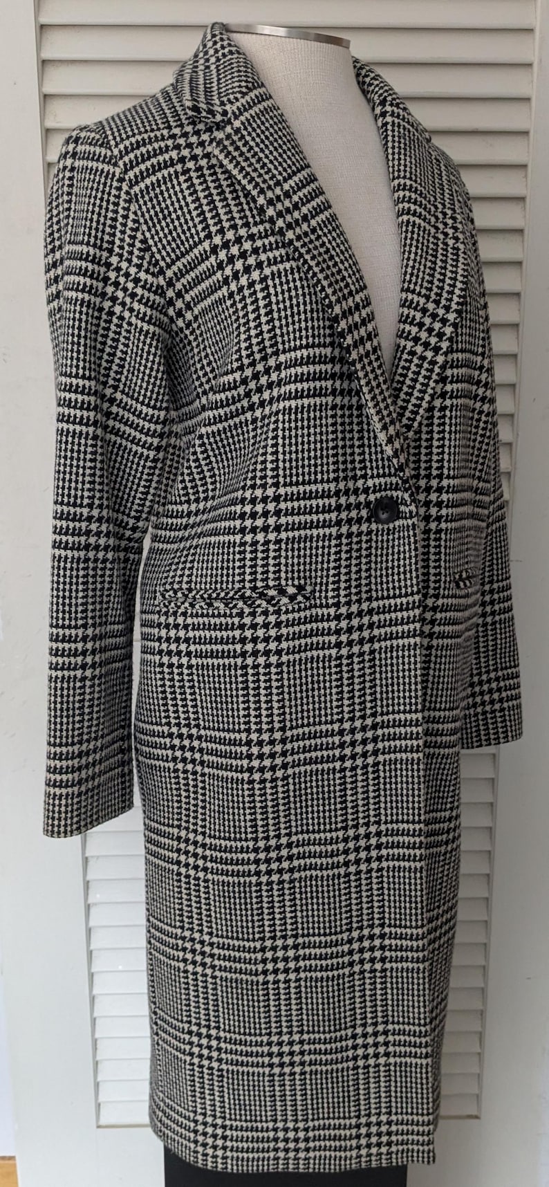 Puede incluir: Un abrigo con estampado de pata de gallo en blanco y negro, con solapas y dos bolsillos delanteros. El abrigo est&aacute; abotonado y tiene mangas largas. El abrigo se exhibe en un maniqu&iacute;.