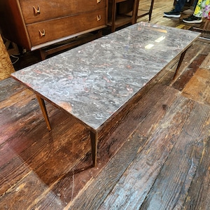 May include: Rectangular coffee table with a gray and red marble-patterned top. The table has four slender, angled wooden legs. The table is approximately 60 inches long and 20 inches wide.