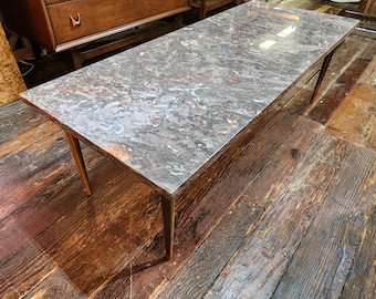 May include: Rectangular coffee table with a gray and red marble-patterned top. The table has four slender, angled wooden legs. The table is approximately 60 inches long and 20 inches wide.