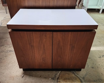 May include: A mid-century modern cabinet with a white top and two wooden doors. The cabinet has a dark brown finish and a black base. The doors have a wood grain pattern. The cabinet is a storage unit.