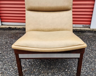 May include: A mid-century modern chair with a light tan seat and backrest. The chair has a dark brown wooden frame with a silver accent. The chair is designed with a low profile and a slightly curved seat.