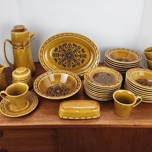 May include: A full vintage mustard-yellow ceramic dinner service. The set includes a coffee pot, creamer, sugar bowl, cups, saucers, bowls, plates, and a serving platter. Each piece features a black floral design.