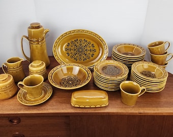 May include: A complete vintage mustard-yellow ceramic dish set. The set includes a coffee pot, creamer, sugar bowl, cups, saucers, bowls, plates, and a serving platter. Each piece features a black floral design.