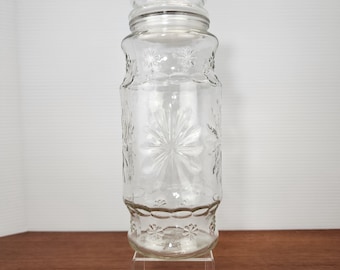 May include: A clear glass jar with a screw-on lid. The jar has a decorative floral pattern and is sitting on a clear acrylic stand. The jar is empty and appears to be vintage.