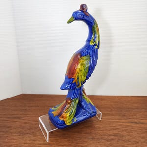 May include: A ceramic peacock figurine in shades of blue, red, yellow, and green. The peacock has a long, curved neck and a detailed tail. The figurine is displayed on a clear acrylic stand.