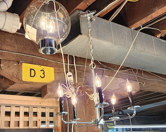 May include: A chrome chandelier with six candle-style lights and black candle sleeves. A large, round glass globe light hangs above. The background includes exposed wooden beams and a yellow sign with the letters "D 3".
