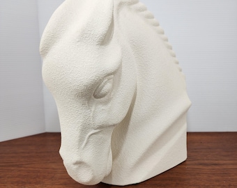 May include: A cream-colored horse head sculpture. The sculpture is a bust, with the head and neck visible. The horse's eye, mane, and facial features are detailed. The sculpture has a textured surface and sits on a wooden surface.