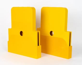 May include: Two bright yellow, geometric-shaped objects with a rectangular base and a rounded top. Each has a circular hole near the top and a vertical slot on one side. The objects are set against a white background.