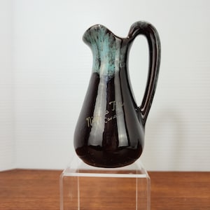 May include: A small, dark brown ceramic pitcher with a teal-colored rim and handle. The pitcher has a glossy finish and the words "Niagara Falls Canada" are written on the side in cursive.