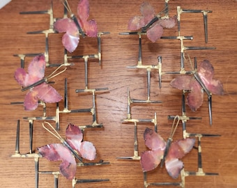 May include: A set of six metal butterfly wall art pieces. Each butterfly has a reddish-pink wing color and is mounted on a black and gold geometric frame. The butterflies are arranged on a wooden surface.