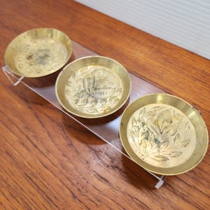 May include: Three small, round, brass dishes with intricate etched floral designs. The dishes are arranged in a row on a clear acrylic stand.