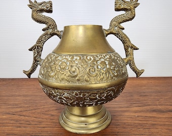 May include: A brass vase with two dragon handles. The vase has a floral pattern around the middle and sits on a round base. The dragons are detailed with scales and claws. The vase is a vintage decorative item.