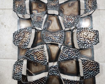 May include: Abstract metal wall art with a geometric design. The artwork features a series of interconnected, textured metal shapes in shades of silver, bronze, and black. The piece is mounted on a rectangular metal backing.