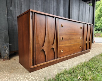 May include: A long wooden sideboard featuring a rich brown finish, three central drawers with small metallic pulls, and two side cabinets with vertical sculpted detailing. The piece has a rectangular top and a solid base, showcasing wood grain.