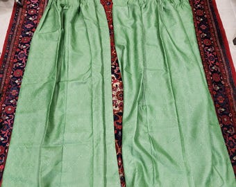 May include: Two light green, patterned curtains are laid out on a patterned rug. The curtains are made of a textured fabric and are folded. The curtains are a solid color and have a subtle floral pattern.