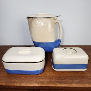 May include: A set of three vintage ceramic kitchenware pieces in white with blue stripes. The set includes a pitcher, a butter dish, and a covered container.
