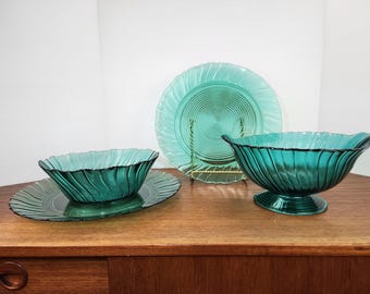May include: A set of teal-colored glass serving dishes. The set includes a large plate, a serving bowl, and a smaller oval-shaped dish. Each piece has a ribbed design and a scalloped edge. The dishes are displayed on a wooden surface.