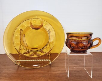 May include: Amber-colored glass teacup and saucer set. The teacup has a rounded shape with a handle and a pattern of raised circles. The saucer set includes a large plate, a smaller plate, and a small, round dish. The set is displayed on a wooden surface.