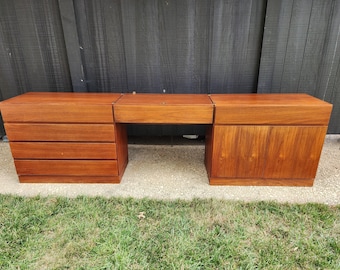 May include: A long, rectangular wooden vanity desk featuring a warm brown finish. The left side consists of a stack of four horizontal drawers, while the right side features a cabinet with two doors. A central flat surface connects the two sections.