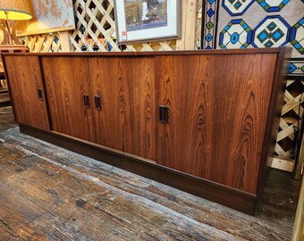 May include: A long, rectangular wooden sideboard featuring a rich, dark brown wood grain pattern. The piece includes four sliding cabinet doors, each equipped with vertical, dark-toned metal handles, and it rests on a solid, recessed base.