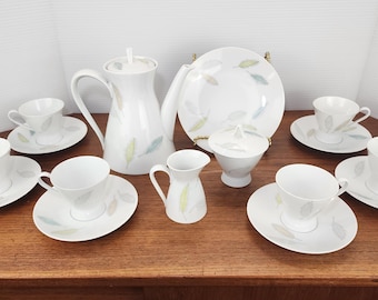 May include: A white porcelain tea set with a leaf design in pastel colors. The set includes teacups, saucers, a teapot, a creamer, a sugar bowl, and a serving plate. The set is arranged on a wooden surface.