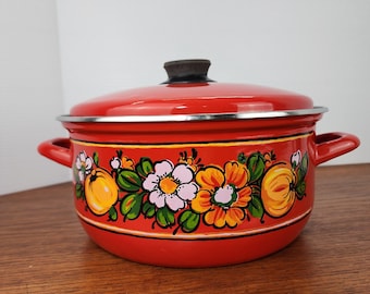 May include: A red enamel pot with a lid and two handles. The pot is decorated with a floral design featuring white, orange, and yellow flowers, and yellow fruit. The pot is sitting on a wooden surface.