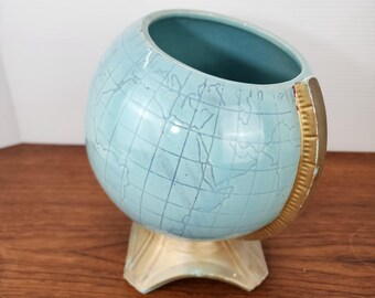 May include: A light blue ceramic globe-shaped planter with a gold-colored band and base. The globe features a map design with continents and grid lines. The interior is a darker shade of blue.