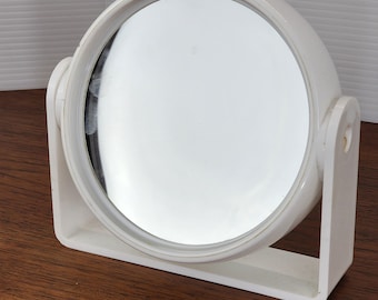 May include: A white, round vanity mirror with a circular frame and a rectangular base. The mirror is supported by two white brackets on either side. The mirror is set on a wooden surface.