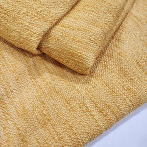 May include: Close-up of folded, textured fabric in a warm yellow hue. The material has a woven look with a slightly raised, nubby texture. The folds create shadows, emphasising the fabric's depth and detail. The fabric is likely for clothing or home decor.