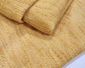 May include: Close-up of a folded piece of textured fabric in a warm yellow color. The fabric has a woven appearance with a slightly raised, nubby texture. The folds create shadows, highlighting the fabric's depth and detail.