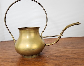 May include: A vintage brass watering can with a curved spout and a large, arched handle. The can has a rounded body and a small, circular base. The brass has a warm, golden hue and shows signs of age.