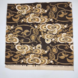 May include: A piece of fabric with a repeating pattern of stylized floral and leaf motifs in shades of brown, beige, and cream against a dark brown background. The fabric has text along the bottom edge.