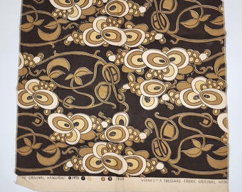May include: A piece of fabric with a repeating pattern of stylized floral and leaf motifs in shades of brown, beige, and cream against a dark brown background. The fabric has text along the bottom edge.