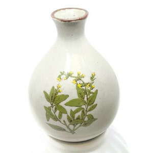 May include: A white ceramic vase with a brown rim and a floral design featuring green leaves and yellow flowers. The vase sits on a white pedestal base.