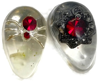 May include: Two clear glass teardrop-shaped pendants with red rhinestone accents. One pendant features a silver spider and the other features a black filigree design.