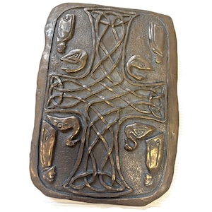 May include: A bronze-colored, rectangular plaque with a Celtic knot design at the center. The plaque features bird-like figures at each corner and along the top and bottom edges. The edges of the plaque have a rough, uneven texture.